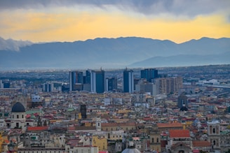 A sprawling urban landscape featuring a large cluster of modern skyscrapers in the background, set against a range of mountains. The foreground is filled with densely packed, older buildings with varied architectural styles and colorful roofs.