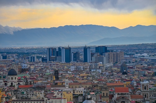 A sprawling urban landscape featuring a large cluster of modern skyscrapers in the background, set against a range of mountains. The foreground is filled with densely packed, older buildings with varied architectural styles and colorful roofs.