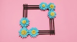 A square-shaped frame made of brown sticks is adorned with blue flowers with yellow centers, set against a pink background.