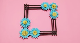 A square-shaped frame made of brown sticks is adorned with blue flowers with yellow centers, set against a pink background.
