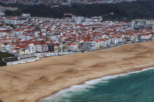 A scenic coastal town with densely packed buildings featuring red-tiled roofs adjacent to a wide sandy beach and a vibrant blue-green ocean. The town is situated at the base of a lush green hillside, offering a picturesque and inviting seaside setting.