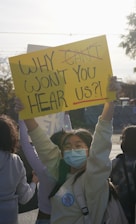 Several people are gathered, with one person prominently holding a yellow sign that reads 'WHY WON'T YOU HEAR US?!'. The person holding the sign is wearing a mask and glasses, and appears to have a badge that says 'NO MORE SILENCE'. The image is backlit by sunlight, creating a dramatic effect.