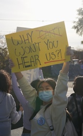 Several people are gathered, with one person prominently holding a yellow sign that reads 'WHY WON'T YOU HEAR US?!'. The person holding the sign is wearing a mask and glasses, and appears to have a badge that says 'NO MORE SILENCE'. The image is backlit by sunlight, creating a dramatic effect.