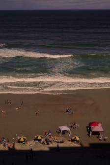 A beach scene with several groups of people enjoying a sunny day. The sandy beach has scattered umbrellas and tents, with a vibrant red-and-white striped canopy standing out. The ocean waves crash gently onto the shore, and there are various beachgoers engaging in leisure activities.