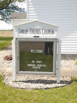 A notice board for Shiloh Friends Church is displayed in a garden setting. The board is encased in a weathered white frame with text indicating a welcome message, Sunday school at 9:30 AM, and a worship service at 10:30 AM. The board is set against a background of a white building and a neatly maintained lawn bordered by gravel.