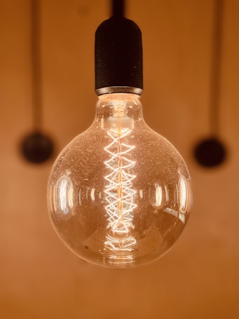 A close-up view of an incandescent light bulb with a glowing filament. The background is warm and blurred, enhancing the focus on the intricate design of the filament inside the transparent glass bulb.