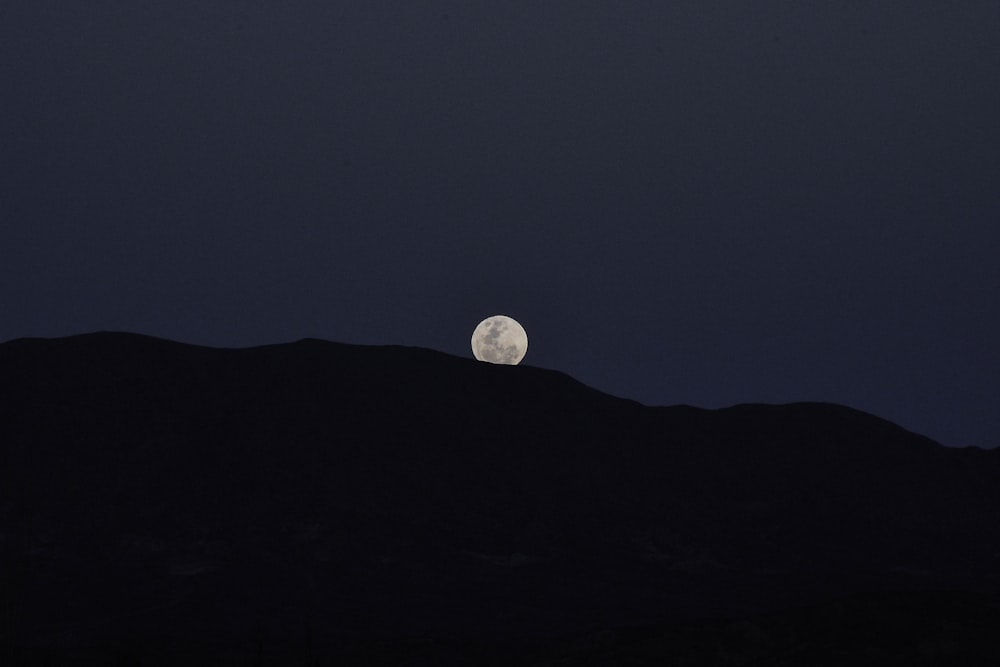 a full moon rising over a mountain with a dark sky