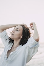 A person with closed eyes, dressed in a white shirt, raises their arms and runs their fingers through their hair. The background suggests a serene and open setting, possibly near the ocean or in an open field.