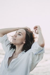 A person with closed eyes, dressed in a white shirt, raises their arms and runs their fingers through their hair. The background suggests a serene and open setting, possibly near the ocean or in an open field.