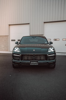 A sleek, black luxury SUV is parked in front of a large garage door. The vehicle features prominent headlights and a shiny exterior, reflecting the surroundings. The setting suggests an industrial or modern garage environment.