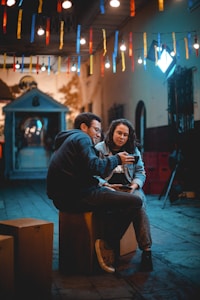 Two people sit on wooden crates in a dimly lit alleyway, illuminated by colorful string lights. They are engaged in conversation, one pointing at something on a phone held by the other. The background shows a quaint building with warm tones, and the setting has a cozy, intimate atmosphere with festive decorations.