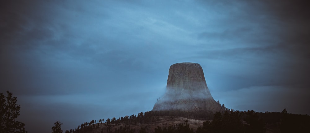 a very tall mountain towering over a forest under a cloudy sky