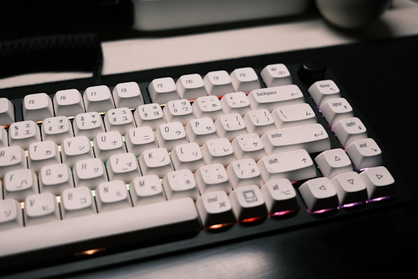 A mechanical keyboard featuring white keycaps with a combination of English and Japanese characters sits on a dark desk. The keys have a prominent, blocky design, and the lighting beneath them emits a subtle glow in various colors including purple and orange.