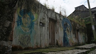 A rustic wall of a building adorned with vibrant street art, featuring two large murals of people with striking colors and abstract elements. The surface of the wall is weathered, with signs of age and vegetation growing on top. The path in front is made of uneven stone steps, leading towards the graffiti.