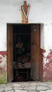 An old, weathered wooden door with peeling paint, partially open to reveal the dimly lit interior of a rustic shop. Inside, various clay pots, bottles, and other items are visible on a table. Over the doorway hangs a wooden cross adorned with green, white, and red ribbons.