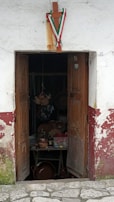 An old, weathered wooden door with peeling paint, partially open to reveal the dimly lit interior of a rustic shop. Inside, various clay pots, bottles, and other items are visible on a table. Over the doorway hangs a wooden cross adorned with green, white, and red ribbons.