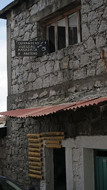 A rustic stone building with a sign advertising services such as masseur and midwife. The building has weathered stone walls and a tin roof overhang. Yellow and black signs next to the entrance list various other services.