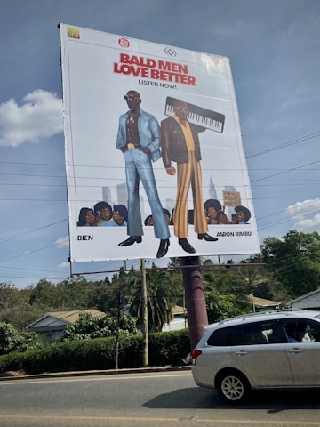 A large street billboard features cartoon illustrations of two fashionable men. The man on the left is wearing a blue suit with a floral shirt and sunglasses, while the man on the right is in a brown jacket with striped pants and holding a keyboard. Below them is text in bold red and black saying 'Bald Men Love Better' and 'Listen Now'. There are small illustrations of audience faces at the bottom. The scene occurs on a clear day with bushes, a house, power lines, and a gray car visible around the billboard.