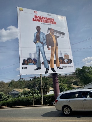 A large street billboard features cartoon illustrations of two fashionable men. The man on the left is wearing a blue suit with a floral shirt and sunglasses, while the man on the right is in a brown jacket with striped pants and holding a keyboard. Below them is text in bold red and black saying 'Bald Men Love Better' and 'Listen Now'. There are small illustrations of audience faces at the bottom. The scene occurs on a clear day with bushes, a house, power lines, and a gray car visible around the billboard.