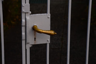 A close-up of a rusty, golden door handle attached to a white metal gate, with a visible keyhole beneath the handle.