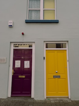 A pair of adjacent doors on a building, one painted a bright purple with a brass mail slot and the other a vivid yellow with a silver mail slot. The wall is a muted gray-green color and a security alarm is mounted above the purple door. A single window with lace curtains is situated above the doors.