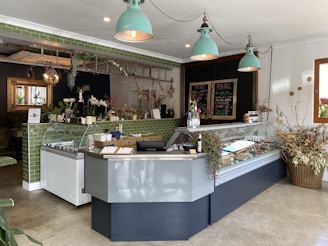 The interior of a stylish café featuring a modern counter with a glass display case containing various food items. Above, there are three teal pendant lights. Green tiled decor adds to the aesthetic, complemented by plants and floral arrangements on the counter and walls. A blackboard menu can be seen, and the overall atmosphere combines rustic and contemporary elements.
