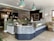 The interior of a stylish café featuring a modern counter with a glass display case containing various food items. Above, there are three teal pendant lights. Green tiled decor adds to the aesthetic, complemented by plants and floral arrangements on the counter and walls. A blackboard menu can be seen, and the overall atmosphere combines rustic and contemporary elements.