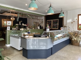 The interior of a stylish caf&eacute; featuring a modern counter with a glass display case containing various food items. Above, there are three teal pendant lights. Green tiled decor adds to the aesthetic, complemented by plants and floral arrangements on the counter and walls. A blackboard menu can be seen, and the overall atmosphere combines rustic and contemporary elements.