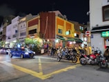 A lively street scene at night featuring a corner with brightly lit restaurants and bars. The exterior of the buildings displays vibrant colors, including orange and yellow. Several people are seated outside, socializing and dining. Parked motorcycles and a blue taxi can be seen on the street, and the area is well-lit with streetlights.