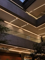 A modern architectural interior featuring multiple levels with sleek lines and glass railings. The scene is illuminated by linear strip lighting embedded in the ceilings, casting a warm glow. Green foliage can be seen peeking from the sides, suggesting an indoor plant or garden area.