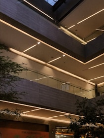A modern architectural interior featuring multiple levels with sleek lines and glass railings. The scene is illuminated by linear strip lighting embedded in the ceilings, casting a warm glow. Green foliage can be seen peeking from the sides, suggesting an indoor plant or garden area.