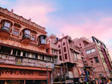 A street scene featuring a row of traditional Indian-style buildings with intricate balconies and carved details. The structures have a pinkish hue, and various signs in local languages are visible, indicating shops and businesses. The sky above is a mixture of blue and pink, suggesting a serene twilight or dawn setting.