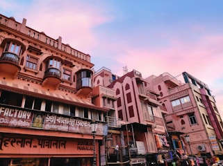 A street scene featuring a row of traditional Indian-style buildings with intricate balconies and carved details. The structures have a pinkish hue, and various signs in local languages are visible, indicating shops and businesses. The sky above is a mixture of blue and pink, suggesting a serene twilight or dawn setting.