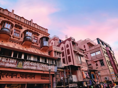 A street scene featuring a row of traditional Indian-style buildings with intricate balconies and carved details. The structures have a pinkish hue, and various signs in local languages are visible, indicating shops and businesses. The sky above is a mixture of blue and pink, suggesting a serene twilight or dawn setting.