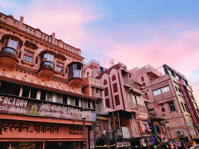 A street scene featuring a row of traditional Indian-style buildings with intricate balconies and carved details. The structures have a pinkish hue, and various signs in local languages are visible, indicating shops and businesses. The sky above is a mixture of blue and pink, suggesting a serene twilight or dawn setting.