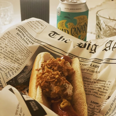 A hot dog topped with mustard and crispy onions is placed on a newspaper-themed paper wrapper. In the background, there's a can of 'The Mad Canary' beverage, and a crystal glass. The setting appears casual and relaxed, likely indicating a meal or snack.
