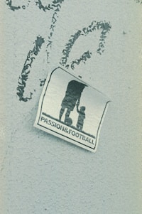 A weathered sticker is affixed to a rough-textured surface, depicting the silhouette of an adult holding hands with a child, next to a soccer ball. The words 'PASSION & FOOTBALL' are printed below the figures, suggesting a connection between family and the sport.