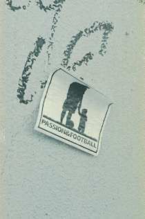 A weathered sticker is affixed to a rough-textured surface, depicting the silhouette of an adult holding hands with a child, next to a soccer ball. The words 'PASSION & FOOTBALL' are printed below the figures, suggesting a connection between family and the sport.