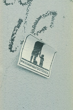 A weathered sticker is affixed to a rough-textured surface, depicting the silhouette of an adult holding hands with a child, next to a soccer ball. The words 'PASSION & FOOTBALL' are printed below the figures, suggesting a connection between family and the sport.