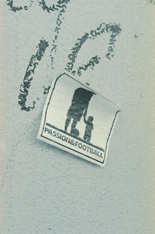 A weathered sticker is affixed to a rough-textured surface, depicting the silhouette of an adult holding hands with a child, next to a soccer ball. The words 'PASSION & FOOTBALL' are printed below the figures, suggesting a connection between family and the sport.