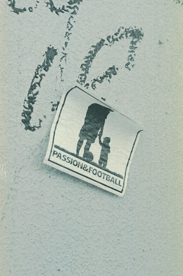 A weathered sticker is affixed to a rough-textured surface, depicting the silhouette of an adult holding hands with a child, next to a soccer ball. The words 'PASSION & FOOTBALL' are printed below the figures, suggesting a connection between family and the sport.