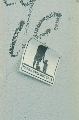 A weathered sticker is affixed to a rough-textured surface, depicting the silhouette of an adult holding hands with a child, next to a soccer ball. The words 'PASSION & FOOTBALL' are printed below the figures, suggesting a connection between family and the sport.