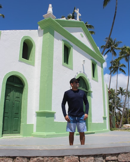 A man wearing a black long-sleeve shirt and blue shorts stands in front of a small church with white walls and light green accents. The church features a cross on the roof and has an arched doorway and windows. Tall palm trees are visible in the background under a clear blue sky.