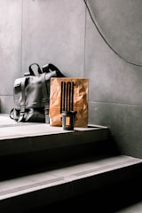 A black leather backpack rests on concrete steps beside a brown paper bag, which has a minimalist design and a takeaway coffee cup in front of it. The setting appears to be indoors with a modern, industrial aesthetic.