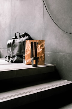 A black leather backpack rests on concrete steps beside a brown paper bag, which has a minimalist design and a takeaway coffee cup in front of it. The setting appears to be indoors with a modern, industrial aesthetic.