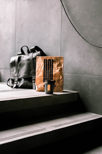 A black leather backpack rests on concrete steps beside a brown paper bag, which has a minimalist design and a takeaway coffee cup in front of it. The setting appears to be indoors with a modern, industrial aesthetic.
