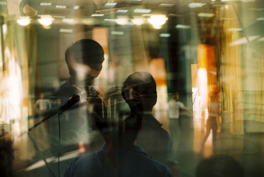 A complex, layered image featuring overlapping silhouettes of people, possibly in a room with lighting fixtures visible. There are reflections and shadows, creating a sense of depth and abstraction.