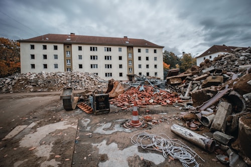 A large, old building with multiple windows stands in the background, surrounded by heaps of construction debris, including piles of bricks, metal scraps, and rocks. Two construction vehicles and a traffic cone are also visible, set against a cloudy sky.