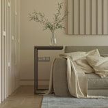 A minimalist living room scene featuring a modern beige sofa adorned with light-colored blankets and pillows. A sleek, rectangular side table holds a clear glass vase with green branches. The room includes light-colored paneling on the walls and a small part of a gray rug visible on the wooden floor.