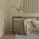 A minimalist living room scene featuring a modern beige sofa adorned with light-colored blankets and pillows. A sleek, rectangular side table holds a clear glass vase with green branches. The room includes light-colored paneling on the walls and a small part of a gray rug visible on the wooden floor.